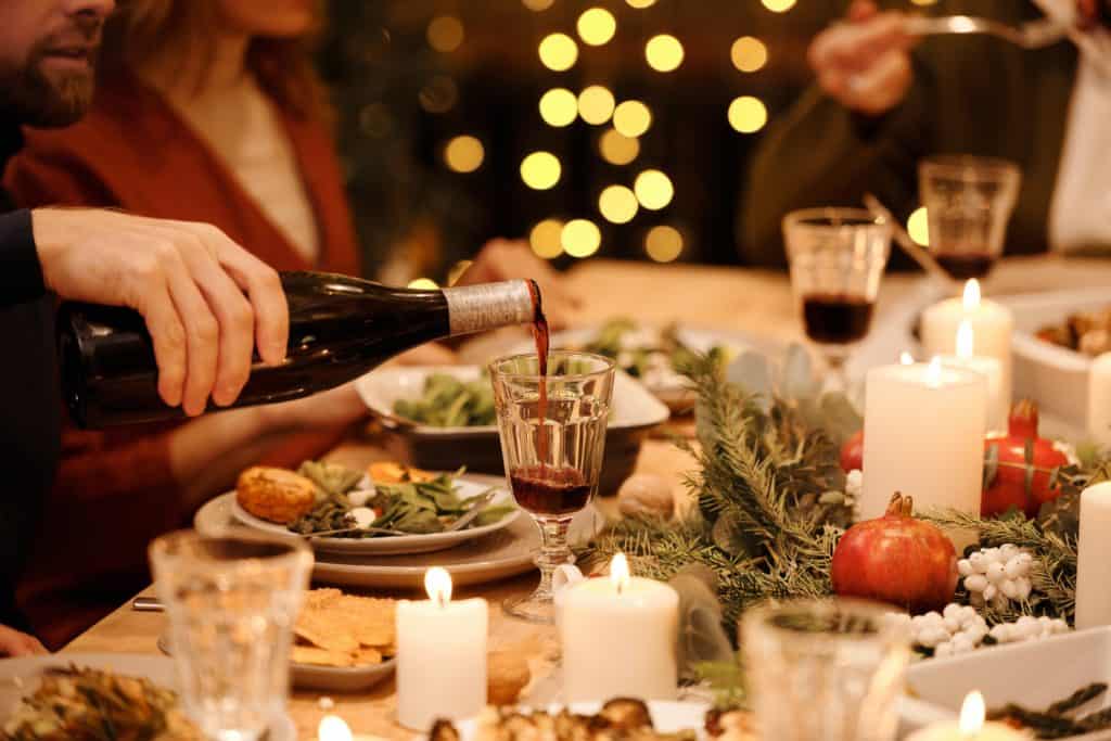 imagem em que uma taça de vidro está em foco com uma mão despejando vinho na mesma. ao redor está uma mesa de natal decorada com velas, ramos de folhas e pratos.