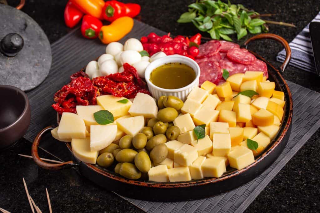 uma tabua de frios com queijos, salames, presunto, azeitonas e um molho
