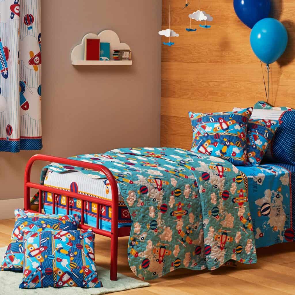 quarto infantil com o tema aviões, o espaço possui janela com cortina, dois balões, uma cama com almofadas, lençóis e kit colcha, e uma prateleira com livros