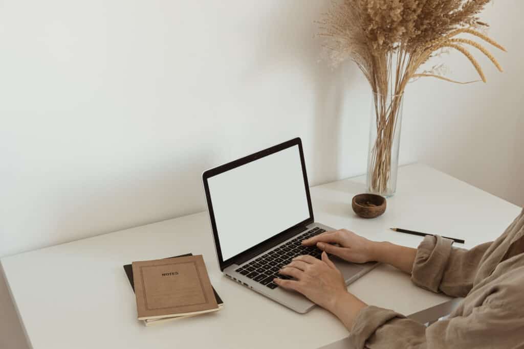 uma mesa de trabalho branca com dois cadernos um lápis e um vaso com plantas, há também um computador sendo utilizado