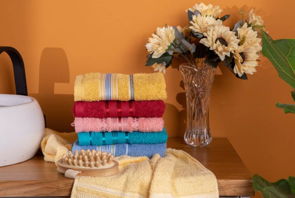 uma pilha de toalhas coloridas acima de uma bancada de banheiro, ao lado um vaso com girassóis e na frente uma escova corporal