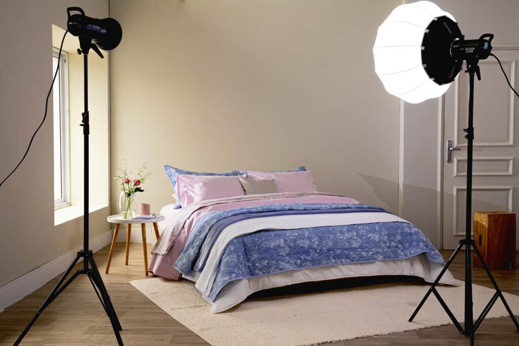 uma cama posta montada em um studio. A roupa de cama é bem completa nos tons de rosa, azul e branco. Ao lado há uma mesa de cabeceira com um vaso com flores e uma caneca, as luminárias do estúdio estão aparentes